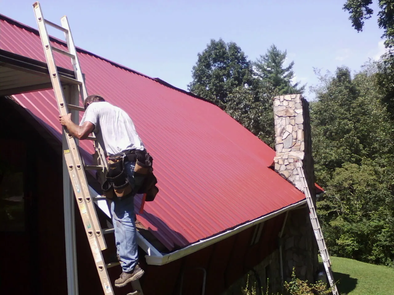 Roofer on a ladder beside a red metal roof and stone chimney — Soffit & Fascia Repair by Roofing Brothers in Barrytown